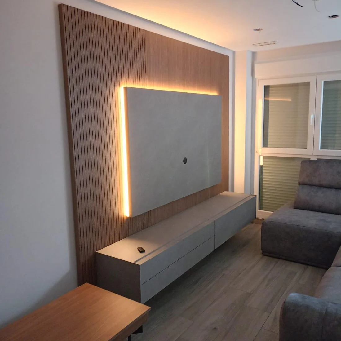 Salón de un cliente acondicionado por completo: panel de televisión a medida en listones de madera con luz indirecta, mueble bajo suspendido, mesa auxiliar y sofá.